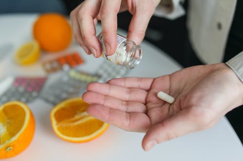 person holding small glass bottle and pill in hands over table with other pills and oranges on it