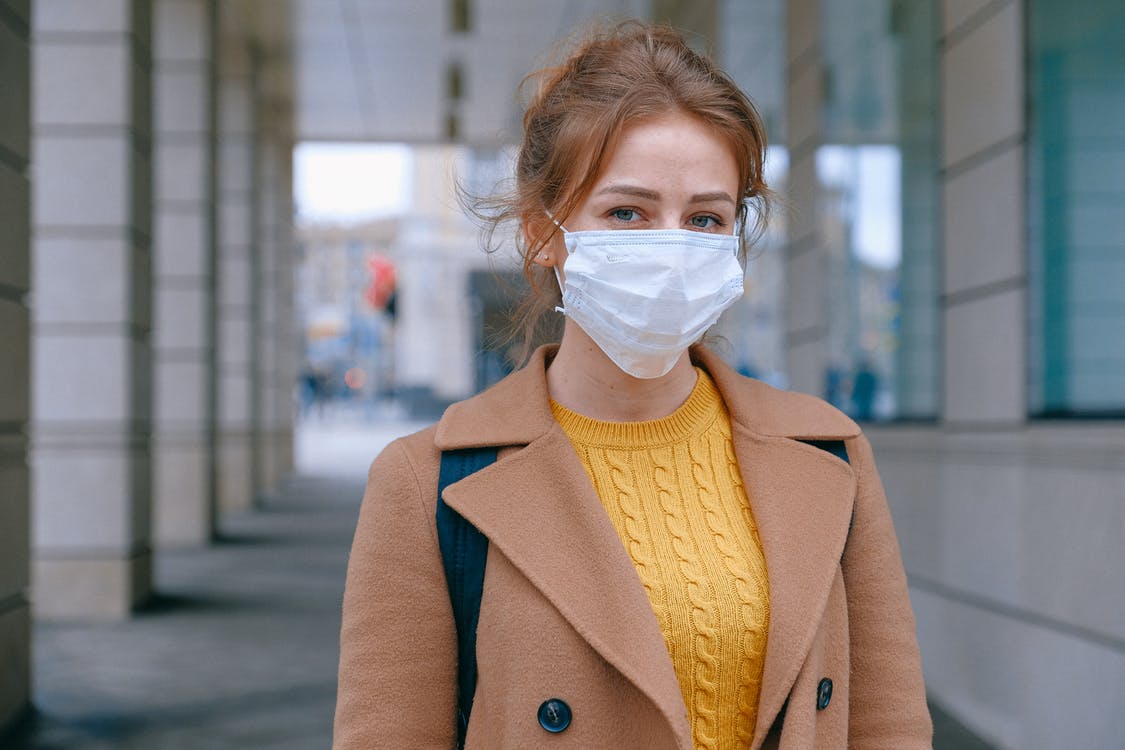 woman looking at camera wearing face mask and brown coat and yellow sweater
