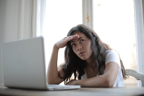 woman with hand on forehead with an irritated expression and sitting at table with laptop open in front of her